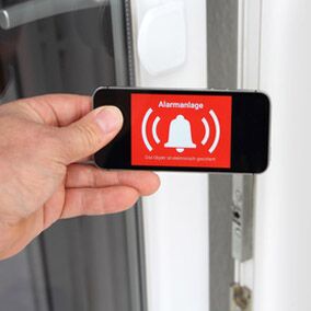 Männliche Hand hält ein Handy auf dem ein Alarmanlagen-Symbol zu sehen ist. Im Hintergrund eine weiße Terrassentür.