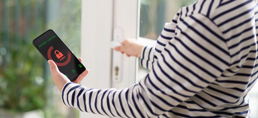 Eine Frau am Fenster mit Hand am Griff richtet das Sicherheitssystem auf ihrem Handy ein