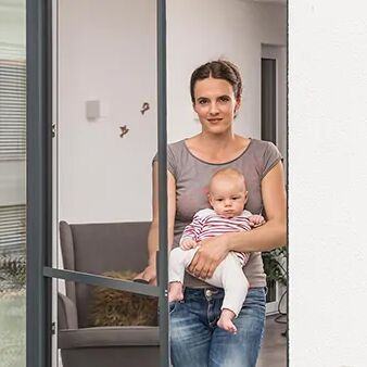 Eine Frau mit Baby auf dem Arm tritt durch eine Terrassentür mit Insektenschutzgitter